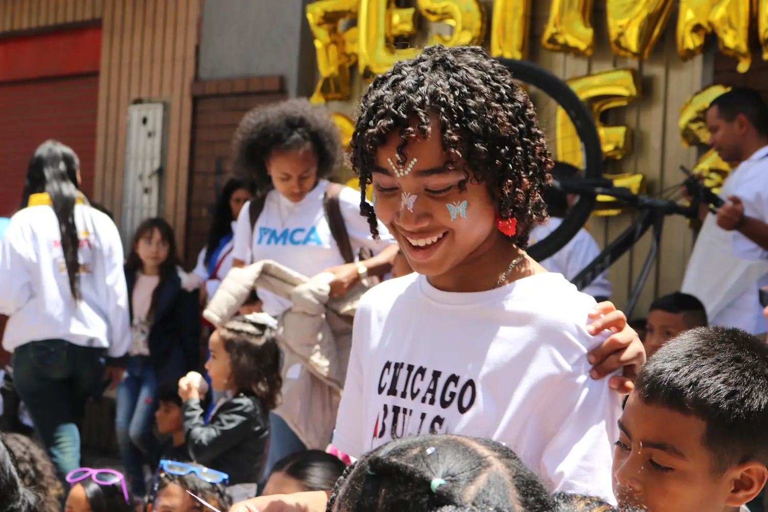 A young participant with butterfly face paint stands among children and community members during the Distintos Somos Todxs street festival in Bogotá, Colombia. People gather in a busy street decorated for the event, which forms part of a project to prevent sexual and gender-based violence supported by Children Change Colombia.