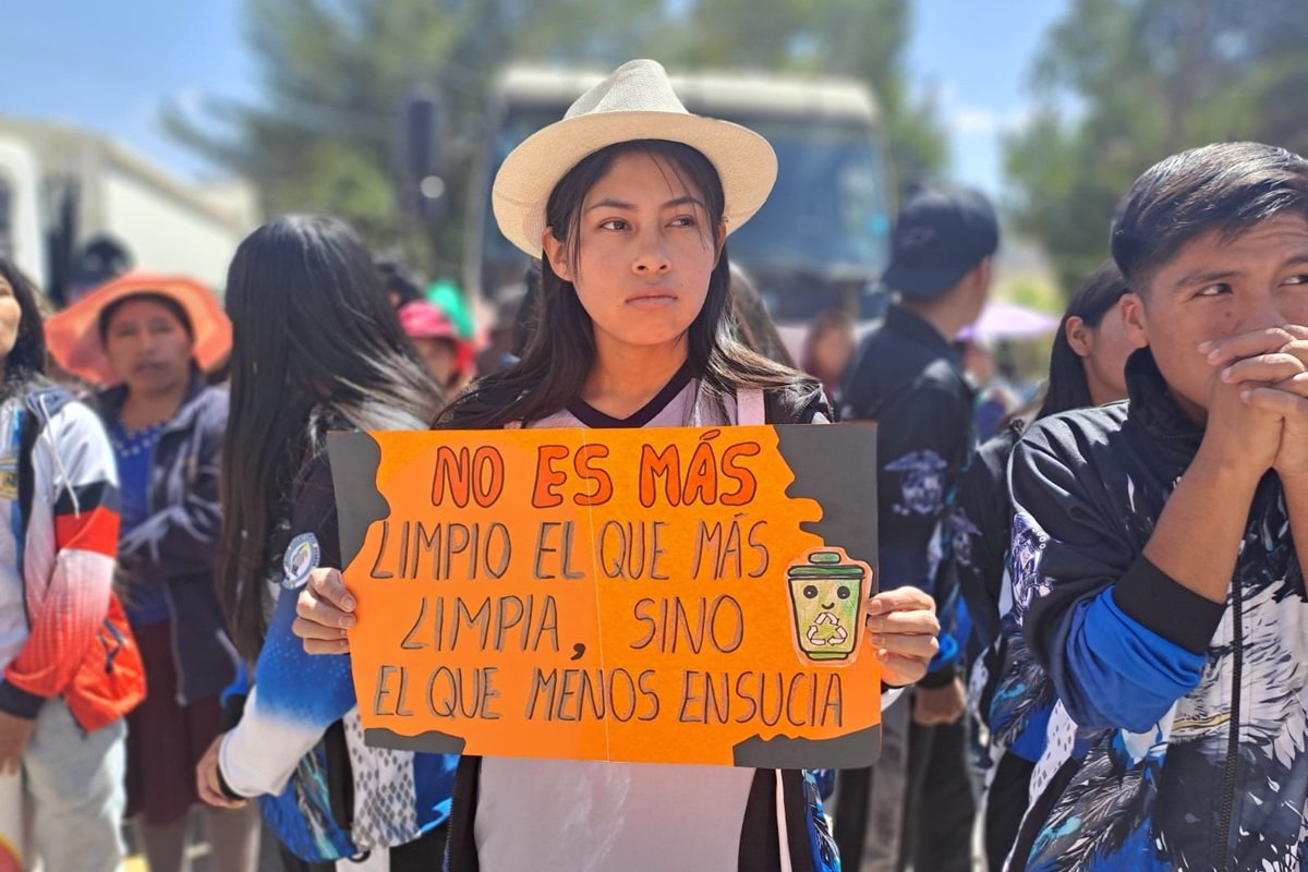 Young People Raising Environmental Awareness in Bolivia | Photo: Plan International | Image provided by Plan International A young participant stands in a crowd during an environmental awareness campaign in Bolivia, holding an orange sign that reads, “No es más limpio el que más limpia, sino el que menos ensucia” (“The cleanest person is not the one who cleans the most, but the one who pollutes the least”). The sign also features a recycling bin symbol promoting environmental responsibility.