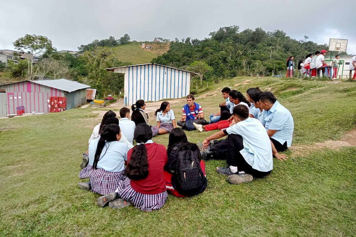 Young people sit in a circle on a grassy hillside during an outdoor workshop in Magdalena Medio, Colombia. A facilitator leads a discussion while participants in school uniforms take part in activities focused on peacebuilding and community dialogue as part of a project implemented by PDPMM with support from CAFOD and the LACT Programme.