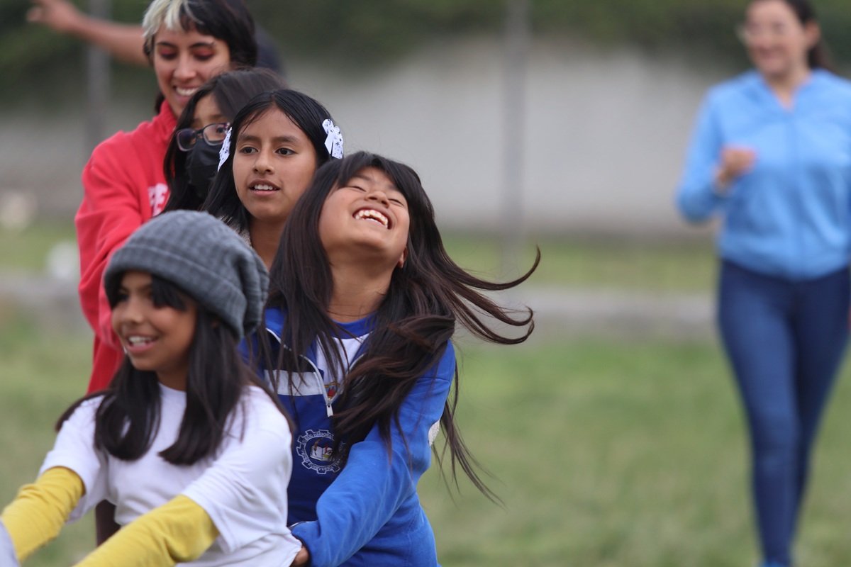 Children participate in a group activity outdoors at the Centro de Apoyo Escolar (CAE) in Ecuador. Several children stand in a line while taking part in a game, with facilitators accompanying the activity in the background. The CAE provides a safe community space where children learn, play and share experiences with one another.