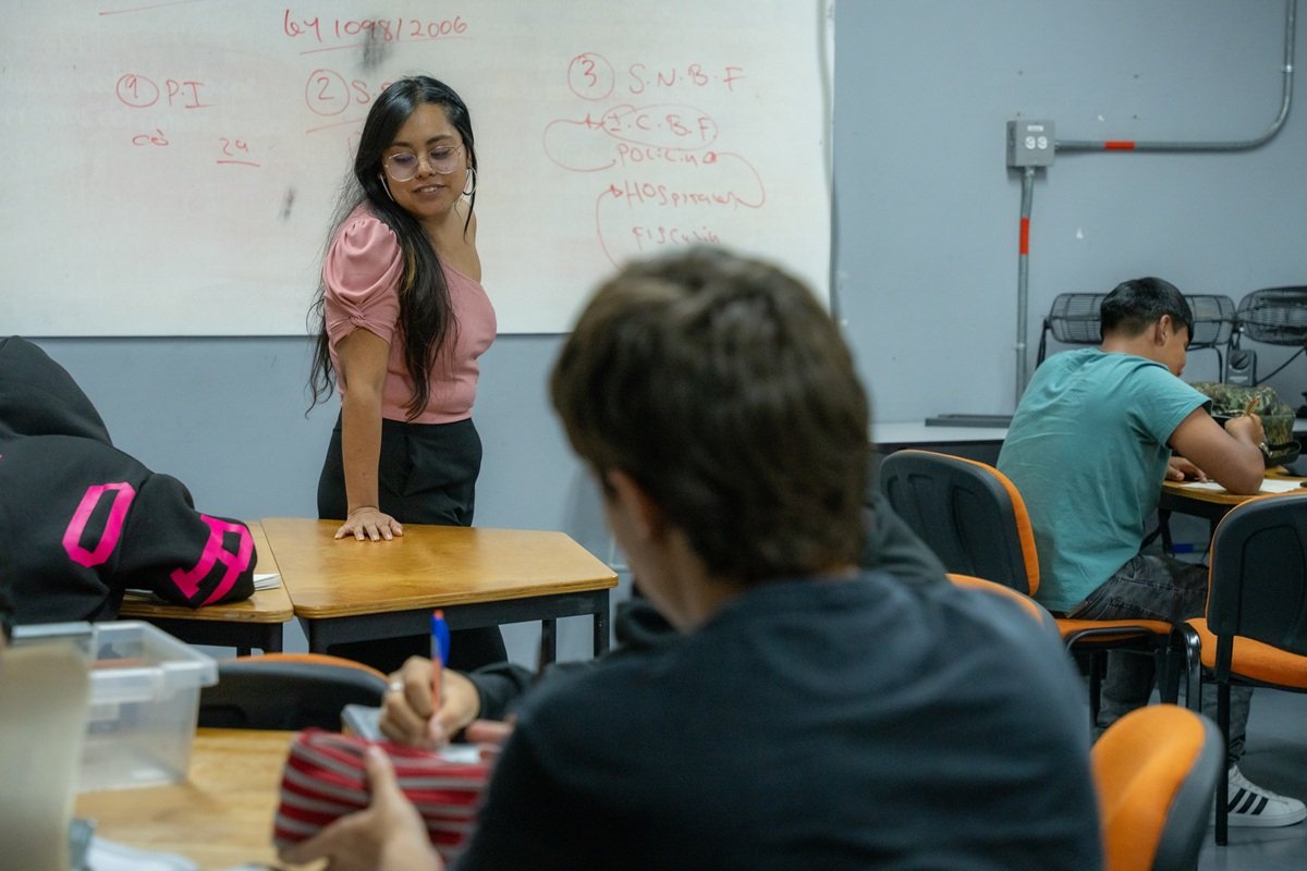 An Enseña por Colombia educator stands at the front of a classroom in Medellín, Colombia, while students work at their desks. A whiteboard with lesson notes is visible behind the educator as students complete classroom activities.
