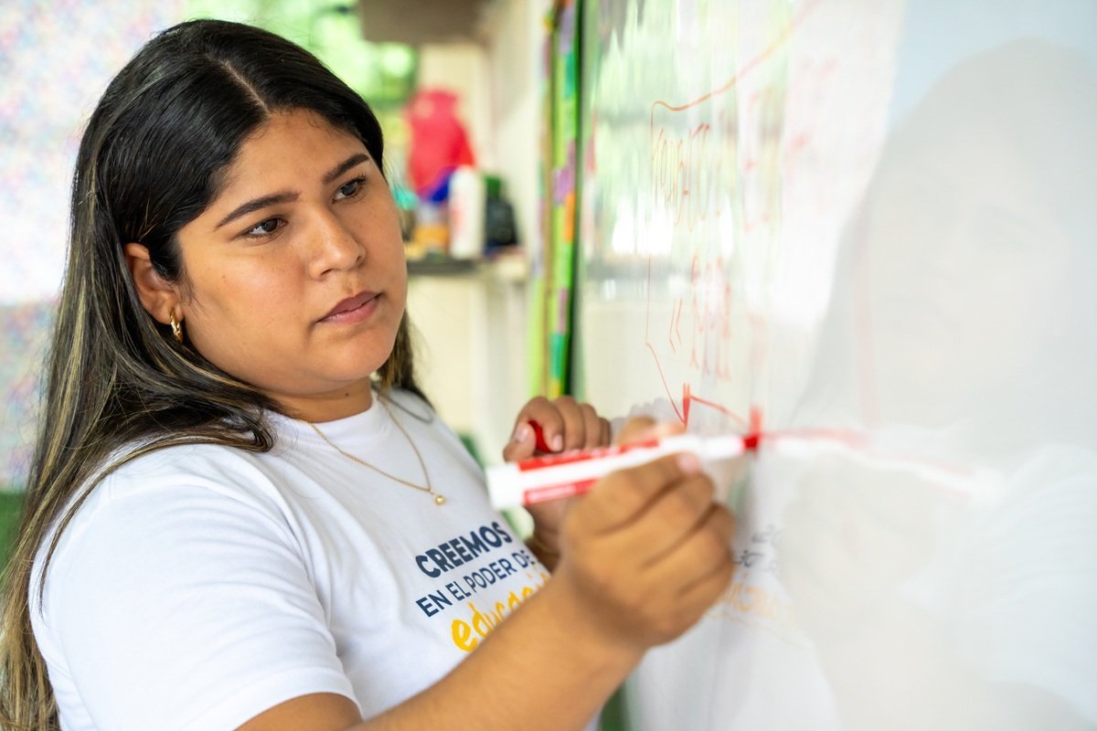 An Enseña por Colombia educator writes on a classroom whiteboard during a lesson in Urabá, Colombia. The educator stands at the front of the classroom while students work at their desks, participating in a learning activity.