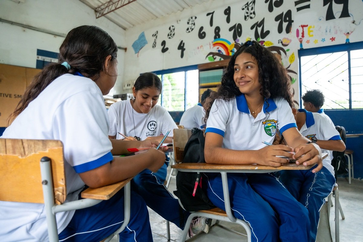 Students sit in small groups inside a classroom in Urabá, Colombia, discussing and working on a learning activity together. Several students hold pens and notebooks while participating in a peer discussion. Colourful educational displays and numbers are visible on the classroom walls.