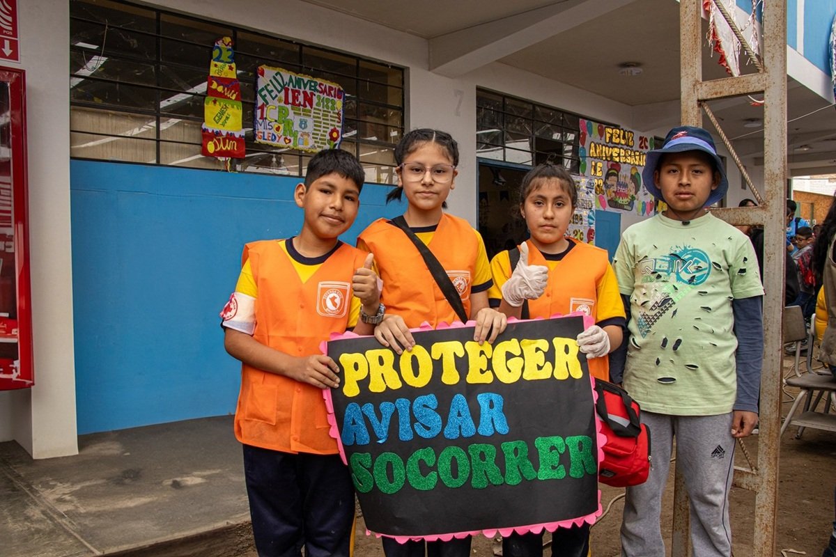 Jóvenes fortaleciendo la preparación para reducir riesgos de desastres en Lima, Perú | Foto: CIDAP | Imagen proporcionada por CAFOD Cuatro participantes jóvenes se encuentran frente a un edificio escolar en Lima, Perú, sosteniendo un cartel con las palabras “Proteger, Avisar, Socorrer”. Tres de ellos llevan chalecos naranjas de respuesta a emergencias y otro participante viste un sombrero azul. La actividad forma parte de una iniciativa de preparación ante desastres implementada por CIDAP con el apoyo de CAFOD y el Programa LACT.