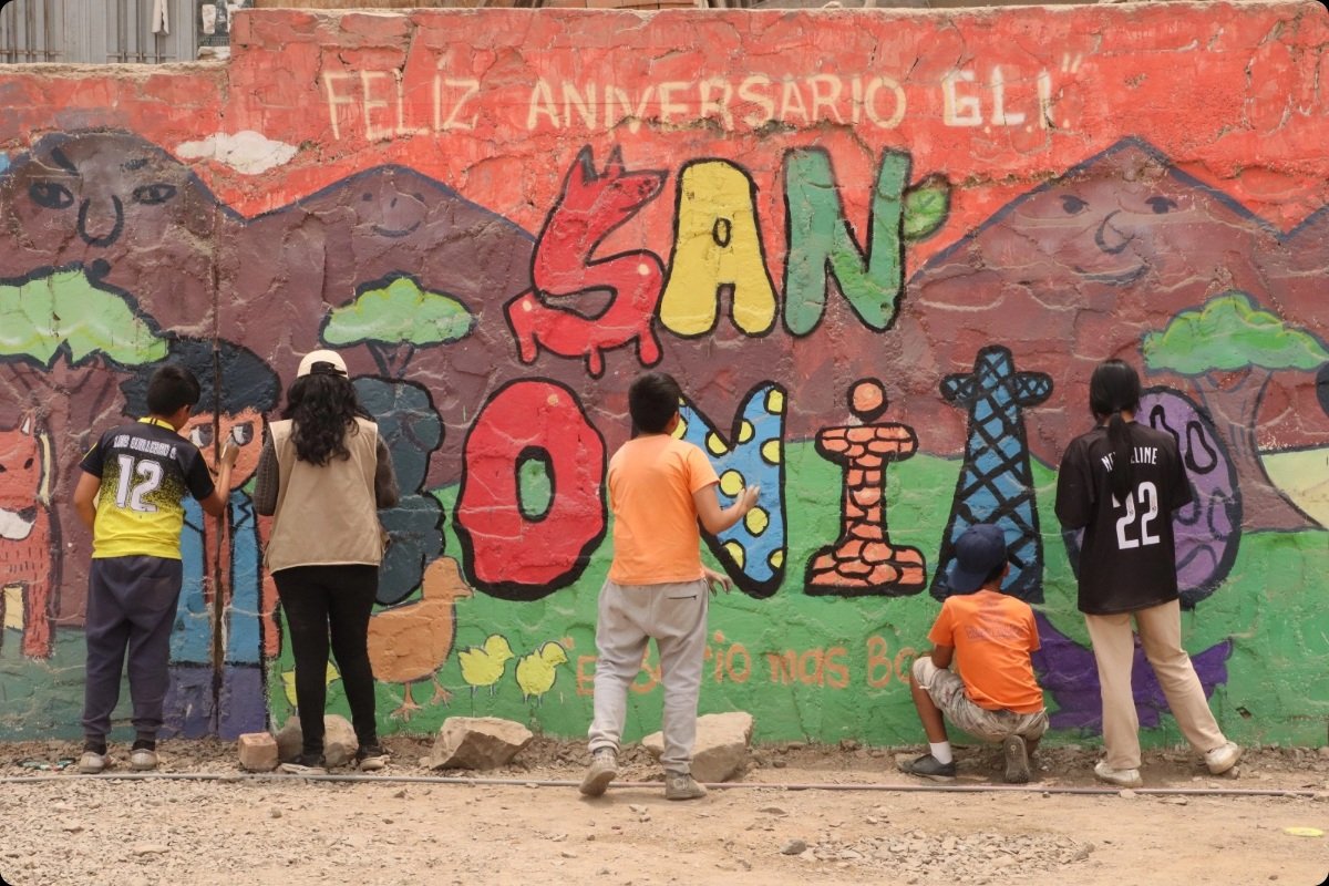 Young people paint a colourful mural on a wall in northern Lima, Peru. The mural features the words “San Boni” alongside drawings of mountains, trees, animals and community symbols. The activity forms part of a project that supports young people to build resilience and defend their rights through creative expression and community engagement.