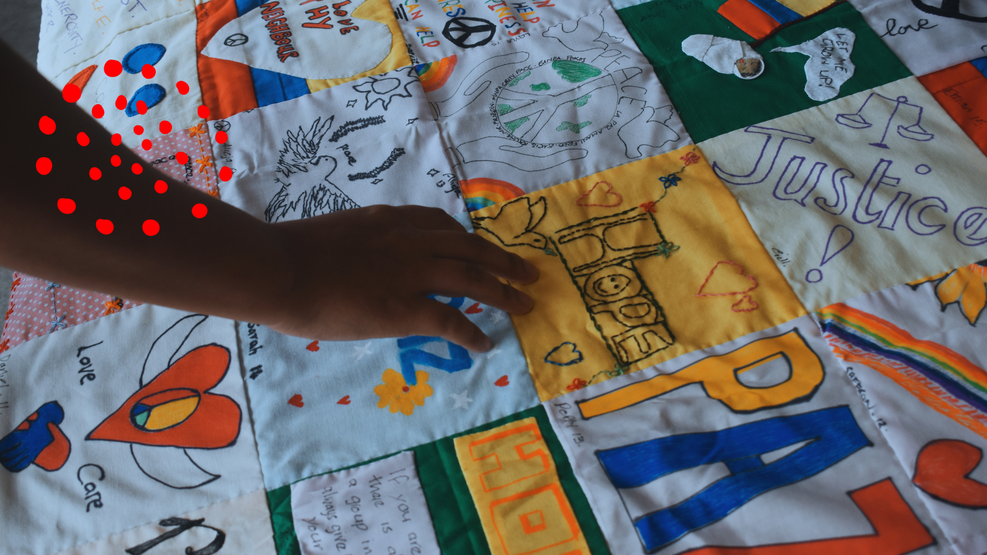 Deseos de niños y niñas para la comunidad de Magdalena Medio en Ecuador. Imagen perteneciente al PDPMM proporcionada por nuestra co-parte CAFOD La mano de un niño descansa sobre un mural de retazos de tela con dibujos y bordados que muestran mensajes y símbolos de paz, justicia, cuidado, amor y comunidad, creados por niños y niñas de Magdalena Medio en Ecuador