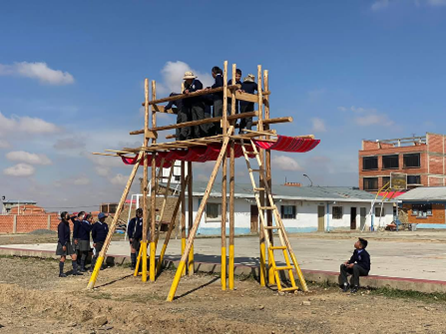 School students gather around and climb a wooden viewing platform in El Alto, Bolivia. Several students stand on the upper observation deck while others watch from below. The structure was built as part of the Green Corridor initiative, creating a community space for environmental education, recreation and outdoor learning.