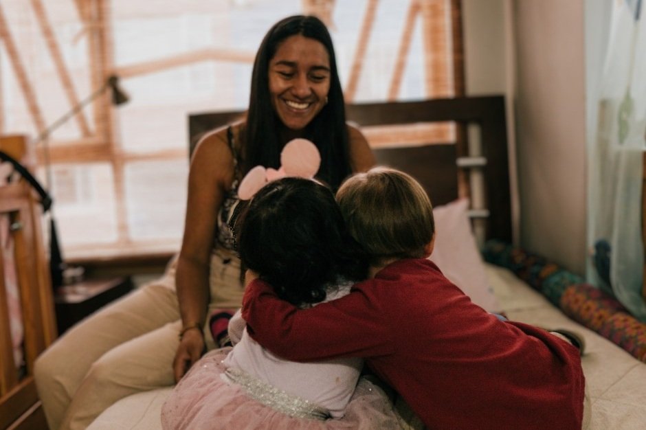 A foster carer sits with two young children on a bed in a family home in Colombia. The children sit closely together while spending time with their caregiver in a domestic setting. The image illustrates the family-based care provided through the Michin foster care programme supported by Lumos Colombia.