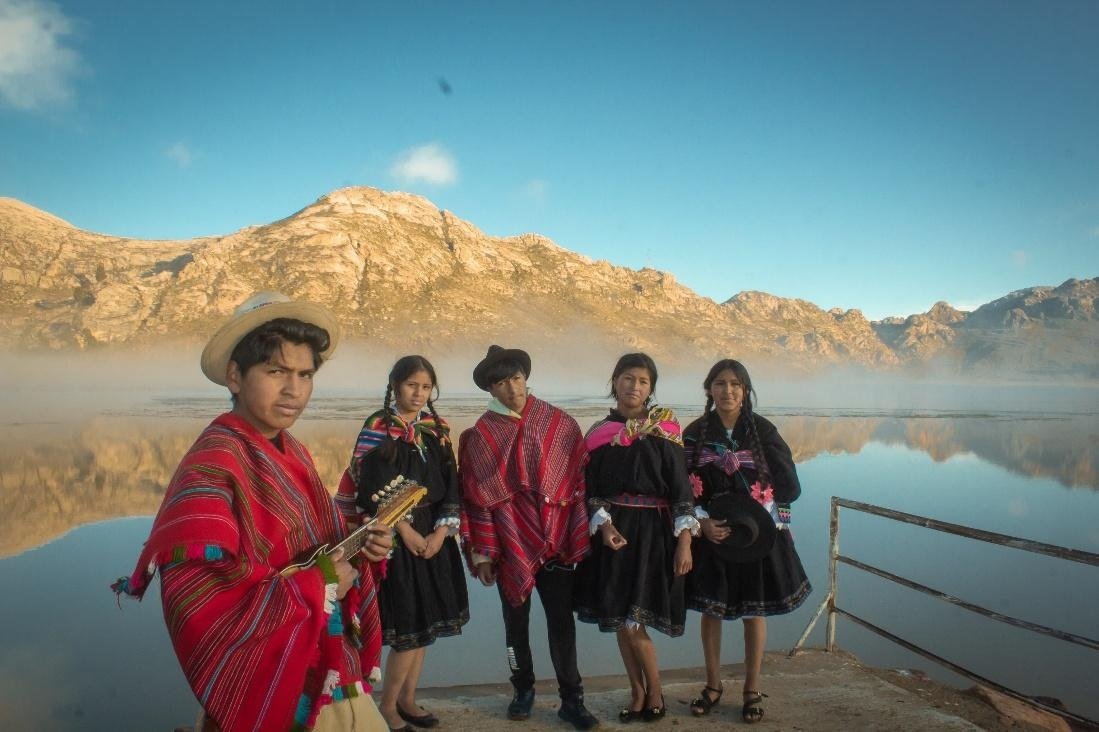 Young People Strengthen Sustainable Community Tourism Skills While Showcasing Cultural Heritage in Bolivia | Photo: Plan International | Image provided by Plan International Participants wearing traditional Andean clothing stand on a lakeside platform in Bolivia, with mountains reflected in the calm water behind them. One participant holds a traditional stringed instrument while the group takes part in a workshop focused on strengthening technical skills for sustainable community tourism.