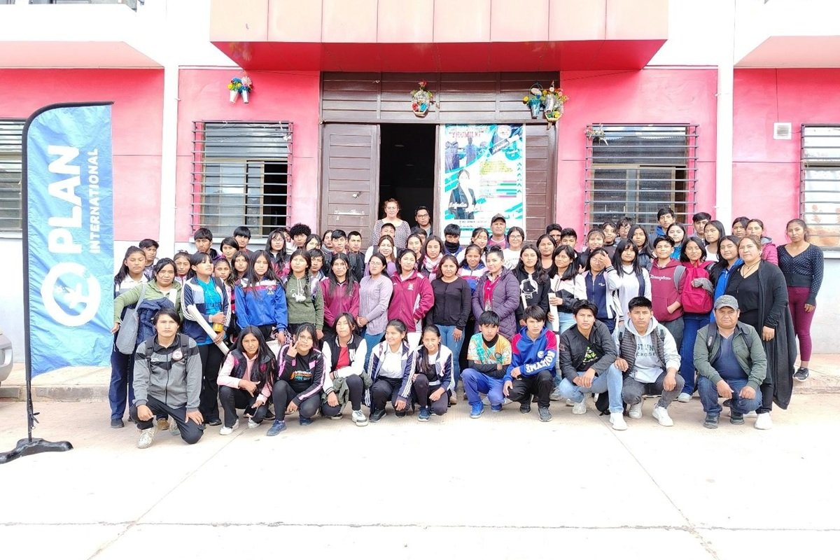 A large group of young people and facilitators gather outside a training venue in Bolivia beside a Plan International banner. The participants have taken part in a workshop focused on entrepreneurship, business management and strategic decision-making for young entrepreneurs.