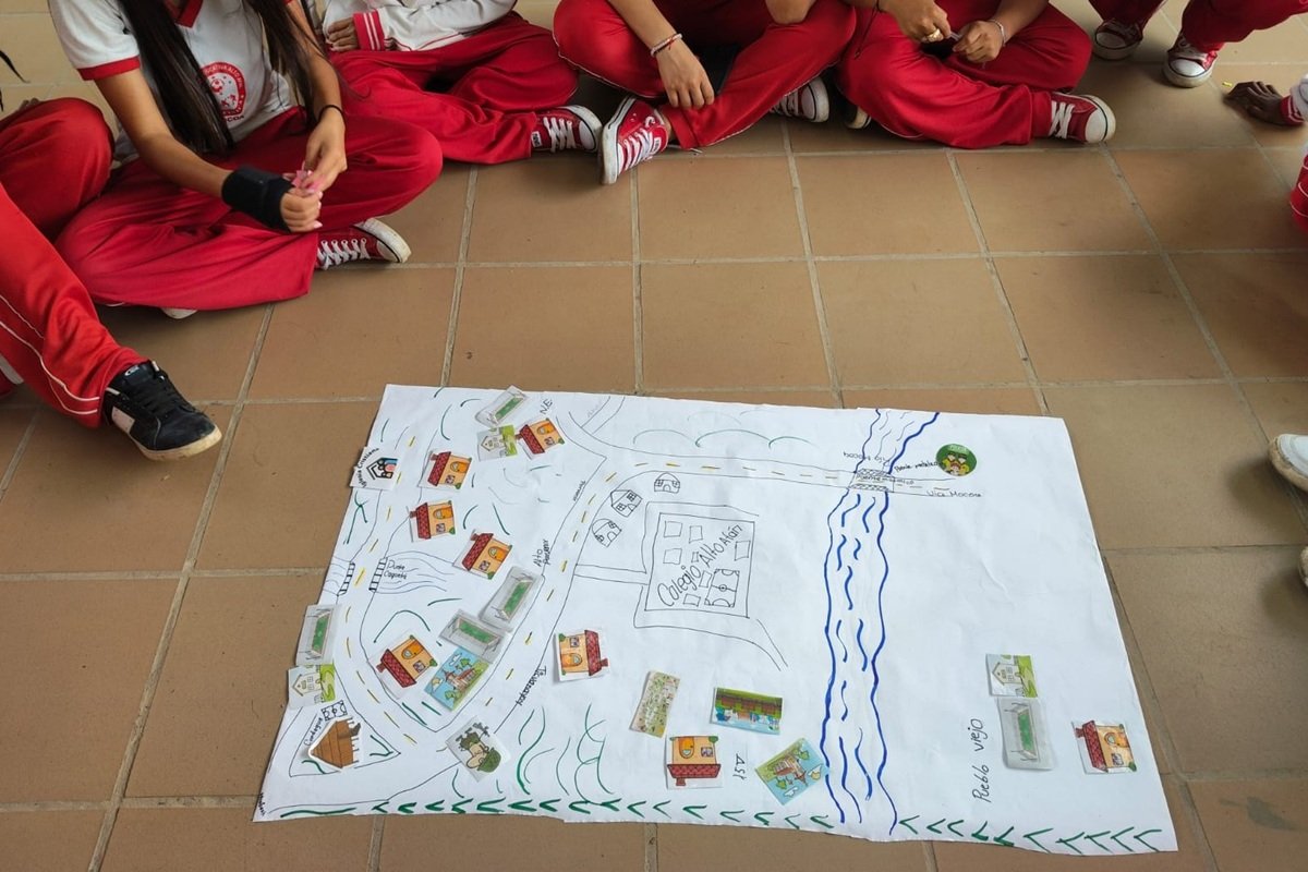 Students sit in a circle around a large hand-drawn community map placed on the floor. The map shows roads, homes, a river and local landmarks represented with drawings and movable pieces. The activity forms part of a participatory risk-mapping exercise where young people identify safety concerns and challenges affecting their community.