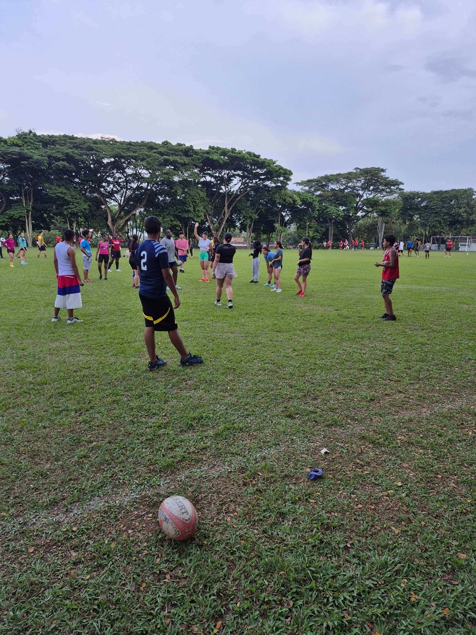 Participants in the Tribu Programme stand on a sports field in Colombia during a training session involving the “Valientes” from the Soroca Programme and students from Pontificia Universidad Javeriana. A rugby ball lies on the grass in the foreground while groups prepare for activities in Ultimate and rugby.