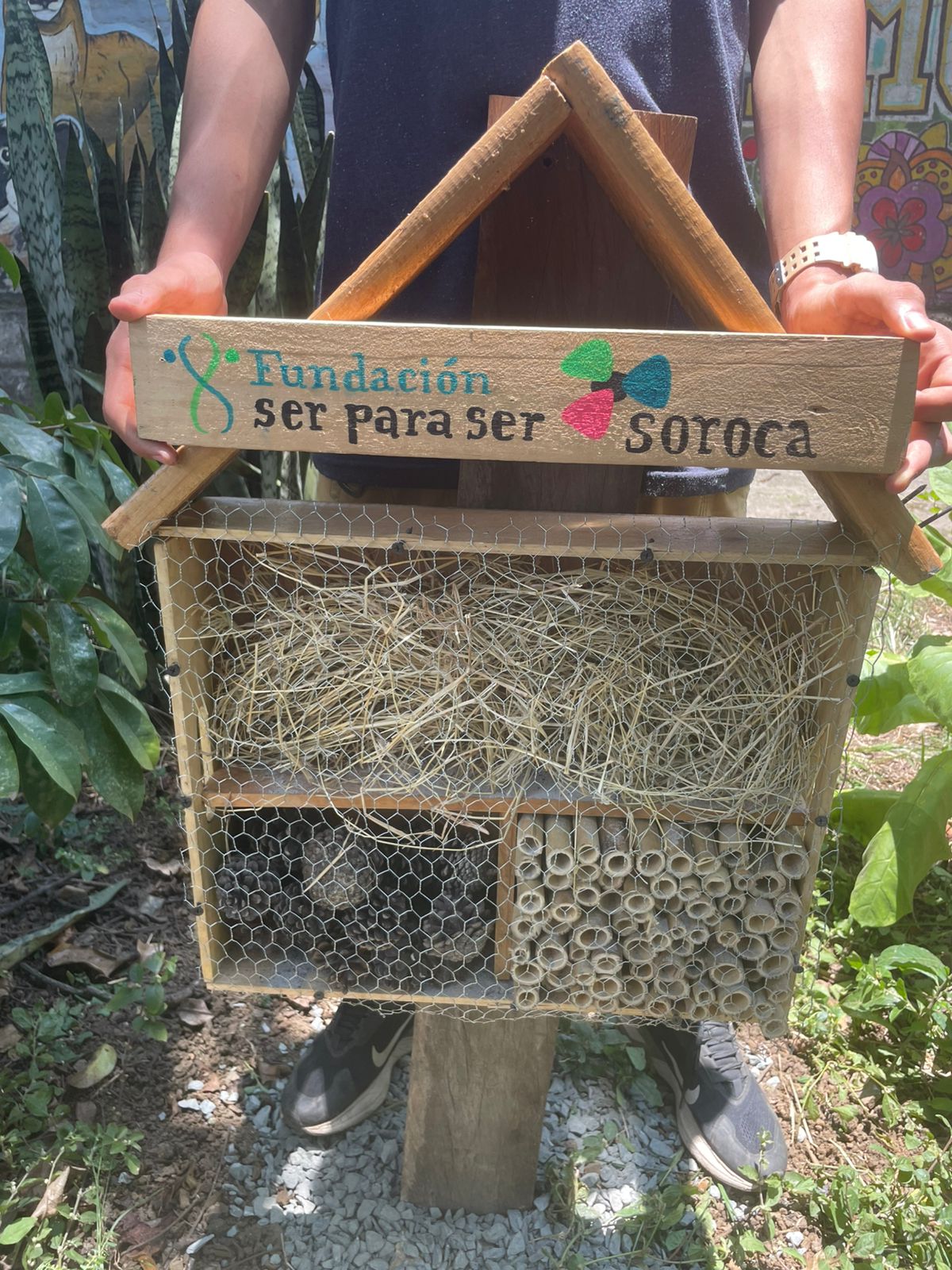 A completed bee and insect hotel stands in a community garden. The wooden structure is filled with bamboo tubes, straw and pinecones that provide habitat for pollinators and other insects. The front displays the names “Fundación Ser Para Ser” and “Soroca.” The hotel was created as part of a community project with the Mi Terracita garden.