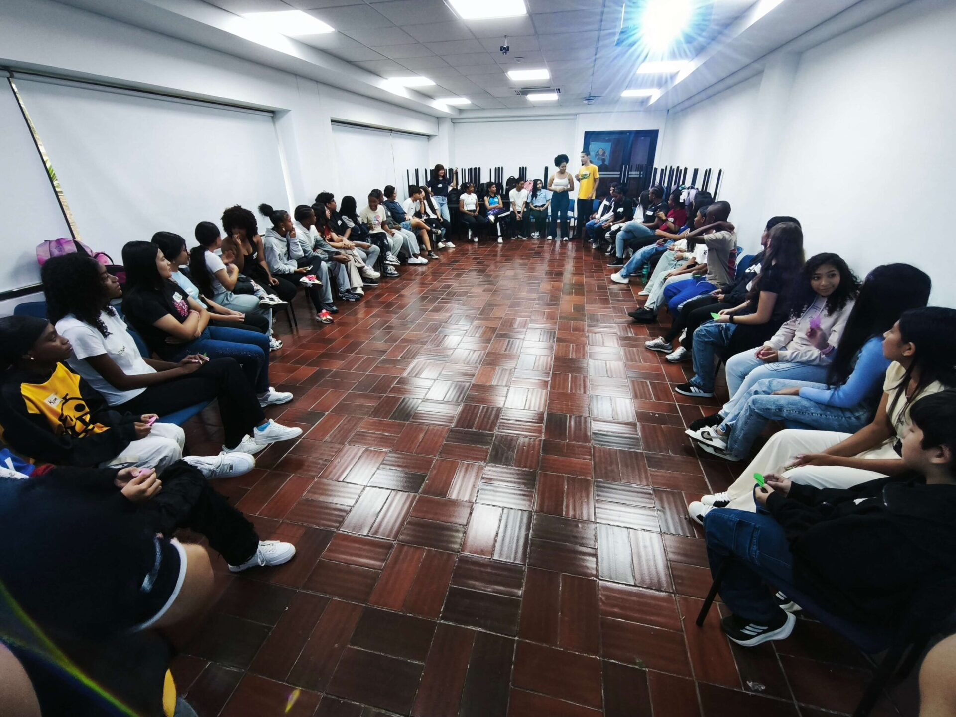 Young participants sit in a large circle during the first regular session of the Soñar level at Pontificia Universidad Javeriana in Colombia. Facilitators stand in the centre of the room while participants take part in a group activity designed to encourage dialogue, participation and peer learning.